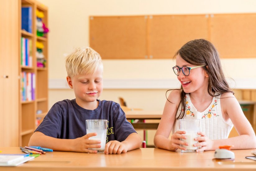 Ein Bub sitzt in der Schulklasse und hält seine Schulmilch in die Kamera.