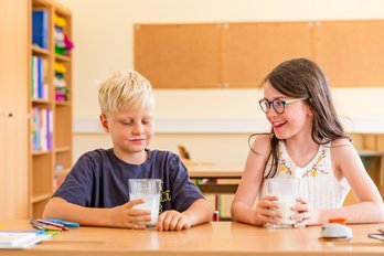 Ein Bub sitzt in der Schulklasse und hält seine Schulmilch in die Kamera.