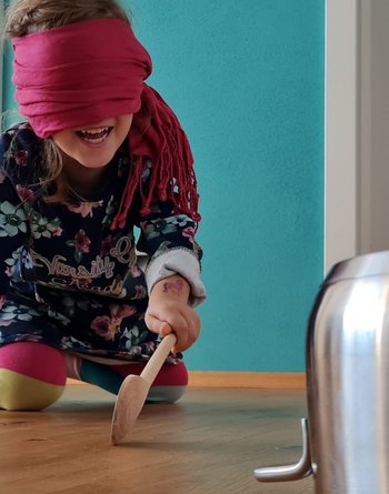 Mädchen hat Augen verbunden und Holzkochlöffel in der Hand und sucht auf Knien den Kochtopf zum Schlagen