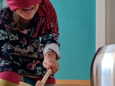 Mädchen hat Augen verbunden und Holzkochlöffel in der Hand und sucht auf Knien den Kochtopf zum Schlagen