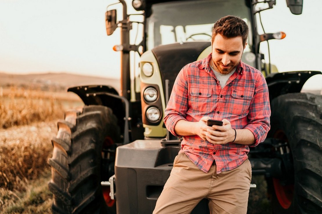 Portrait of smiling farmer using smartphone and tractor at harve