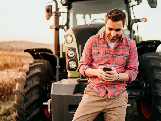 Portrait of smiling farmer using smartphone and tractor at harve