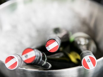 Vier Flaschen liegen im Sektkühler. Auf den Kapseln ist die rot-weiß-rote Weinbanderole mit Adler und Ziffern.