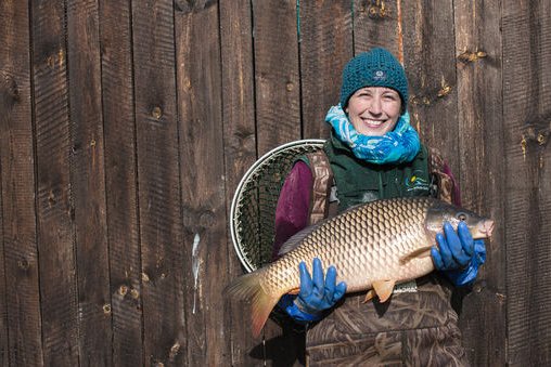 Frau steht im Anglergewand vor Holztor und hält großen Karpfen in den Händen